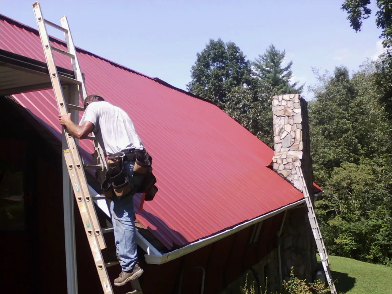 Roofer on a ladder beside a red metal roof and stone chimney — Soffit & Fascia Repair by Roofing Brothers in South Pittsburg